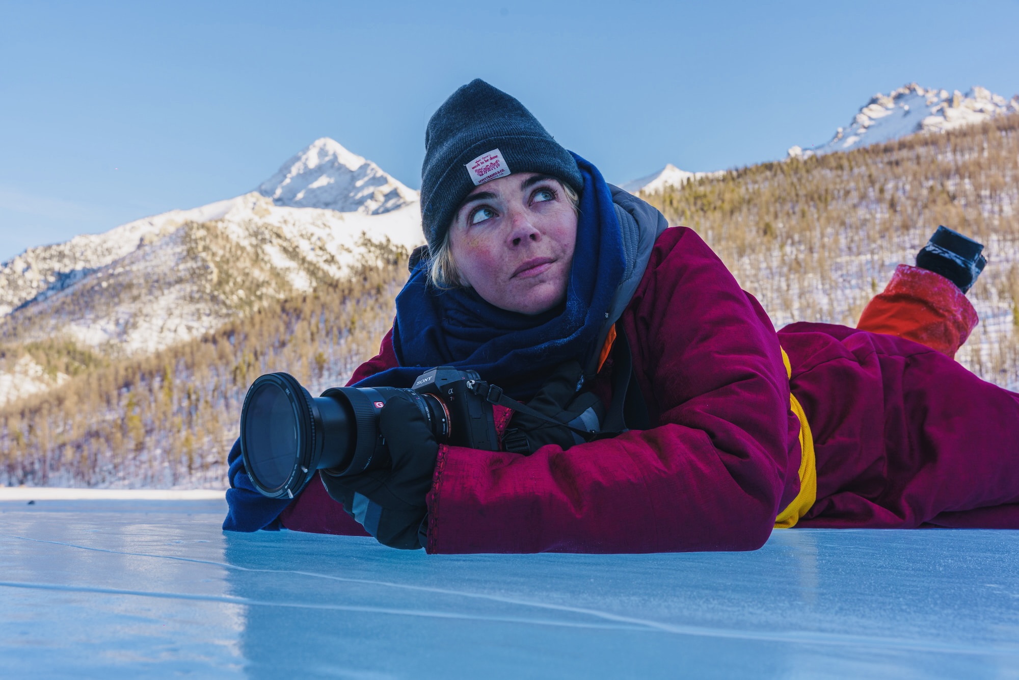 A photographer, in winter boots, lies on the ice to capture Mongolia’s snowy mountain beauty.