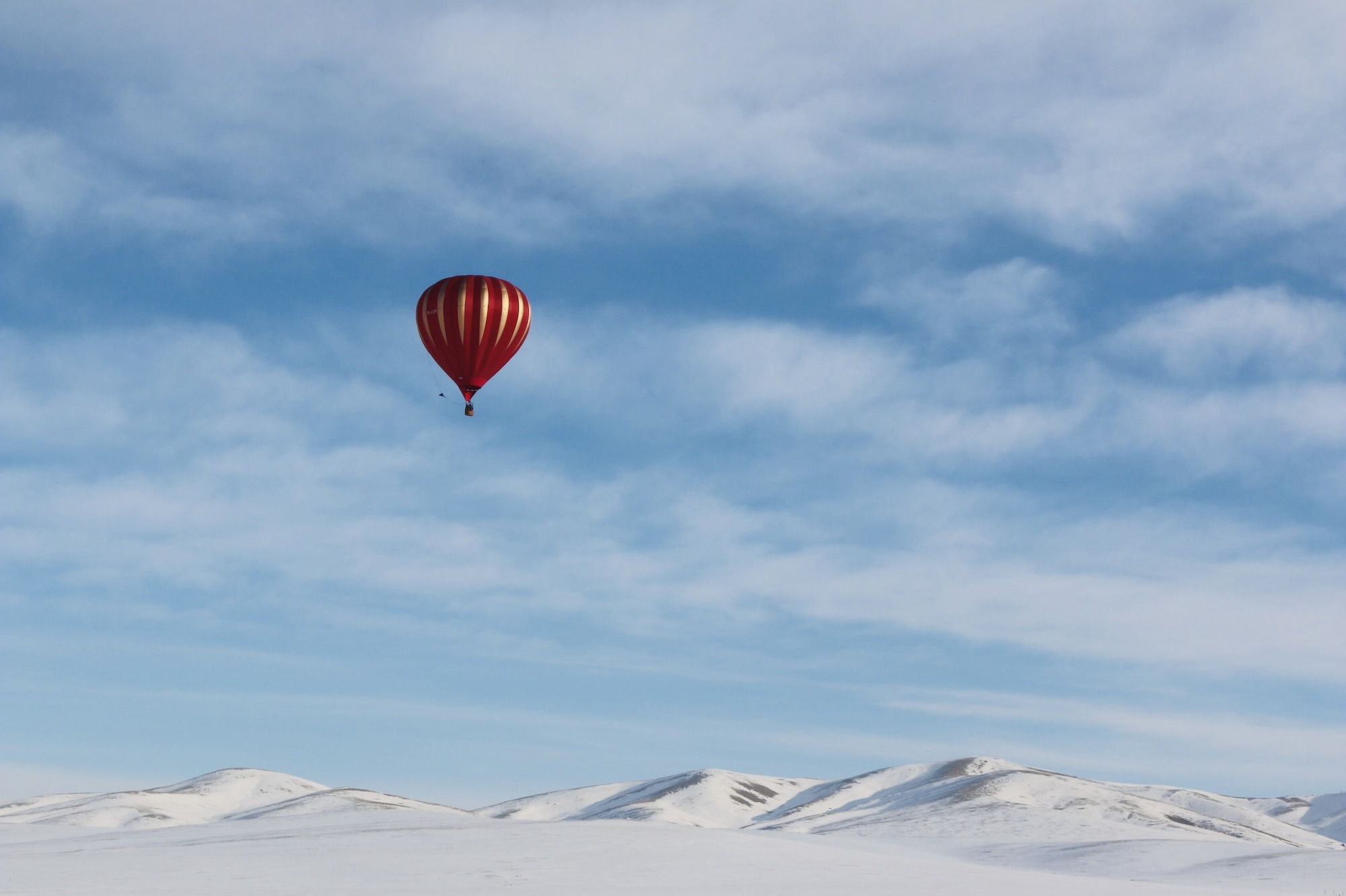 A vibrant hot air balloon drifts over Mongolia’s snowy hills under a eternal  blue sky.